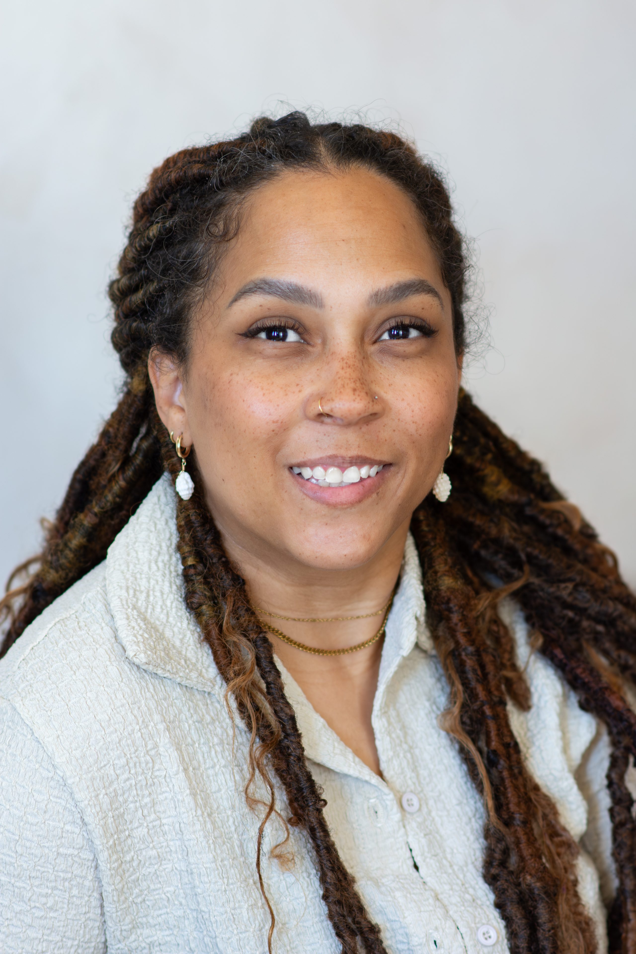 Professional headshot of a smiling staff member with long braided hair wearing a light-colored textured shirt against a neutral background.